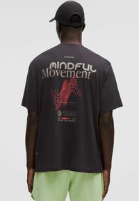 Tricou negru cu mânecă scurtă, având textul "MINDFUL Movement" în roșu și gri, cu un design de model în valuri. Asociat cu pantaloni scurți de culoare verde deschis.