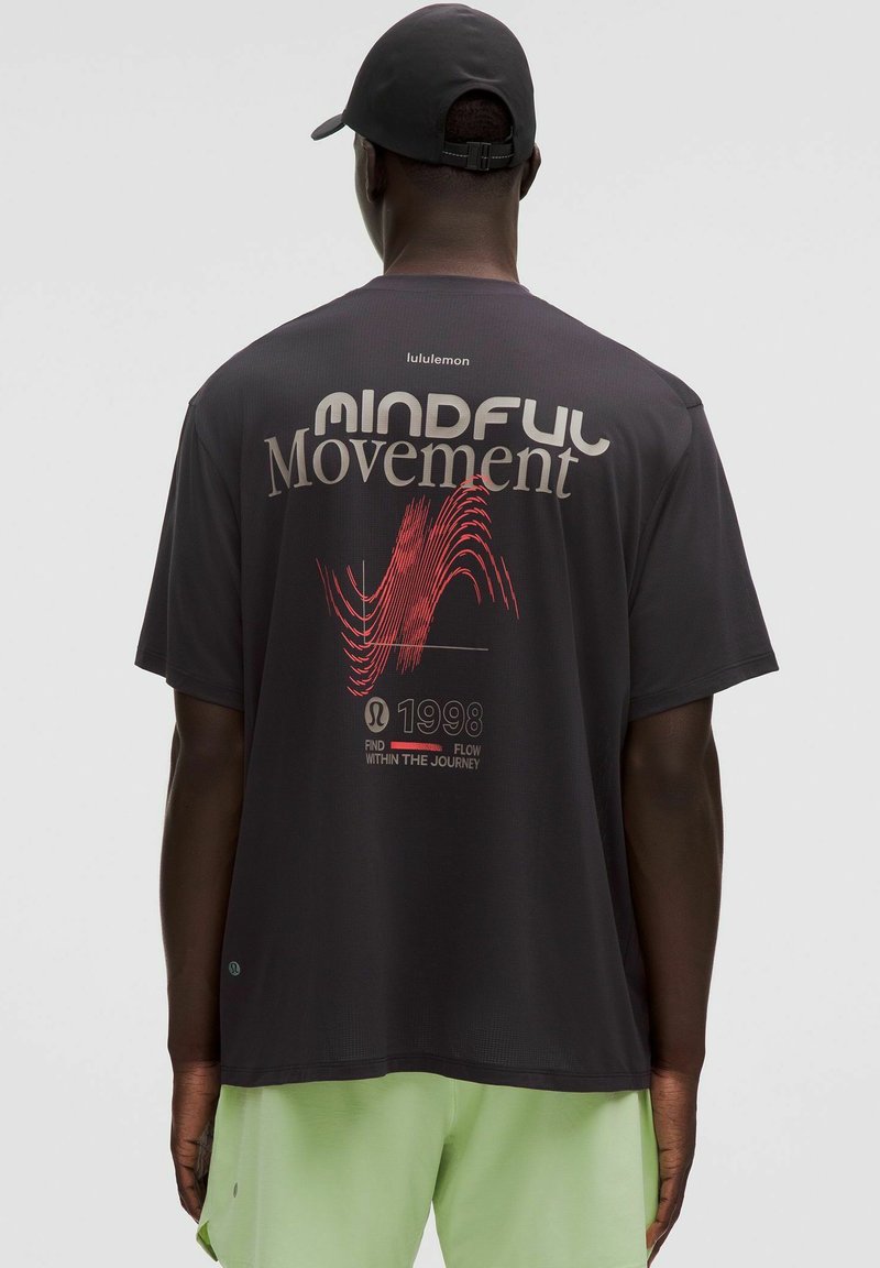 Tricou negru cu mânecă scurtă, având textul "MINDFUL Movement" în roșu și gri, cu un design de model în valuri. Asociat cu pantaloni scurți de culoare verde deschis.