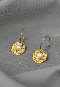 Goldene Hoops tragen strukturierte runde Scheiben mit einem kleinen weißen Perlenakzent, vor einem grauen Stoffhintergrund.