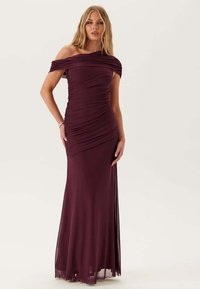 Kvinde iført en gulvlængde, off-shoulder mørk lilla kjole med drapering, der står foran en ensfarvet lyse baggrund.