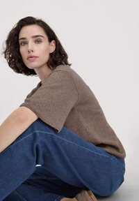Maglione in maglia marrone a maniche corte, abbinato a jeans in denim blu larghi. Lo sfondo è chiaro e neutro, mettendo in risalto le trame dell'outfit.