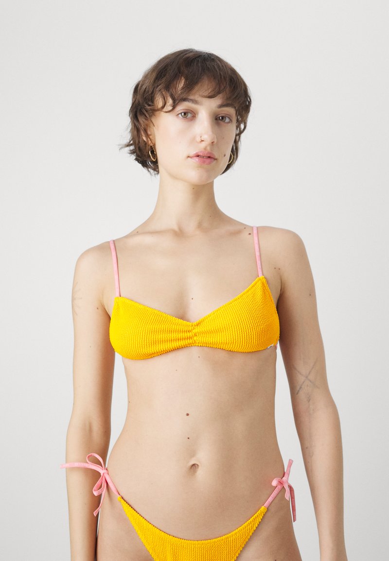 Sárga bordázott bikini felső, rózsaszín pántokkal; az alsó részen oldalt megkötős részletek találhatók. Sima textúra, minimalista dizájn és élénk színkontraszt jellemzi.