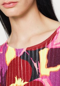Gros plan sur la partie inférieure du visage et le cou d'une personne portant un haut coloré à motif floral avec des tons rouge, jaune, rose et noir.