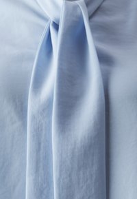 Lyseblå bluse med en drapert knytedetalj ved halsen. Laget av glatt stoff med en myk tekstur, med subtil glans.