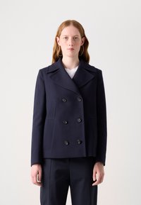 Veste croisée bleu marine avec de grands boutons noirs, revers larges et poches latérales, en tissu structuré. Modèle portant un pantalon noir.