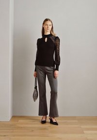 Zwarte top met kantmouwen, sleutelgatdetail bij de halslijn. Grijze uitlopende broeken. Kleine handtas met een patroon, zwarte puntige schoenen. Minimalistische achtergrond.