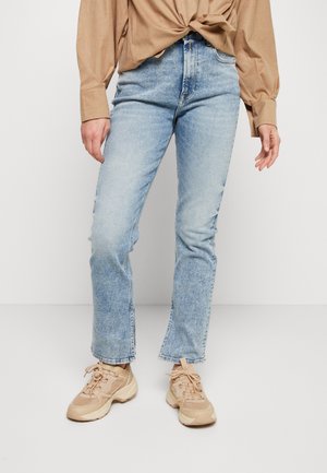 Persoon draagt lichtblauwe rechte jeans, beige chunky sneakers en een beige langarmshirt met een knoopdetail aan de voorkant.