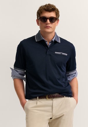 Mann mit dunkler Sonnenbrille, dunkelblauem Poloshirt über gestreiftem Langarmshirt, beigen Hosen und geflochtenem Gürtel, steht mit den Händen in den Taschen.