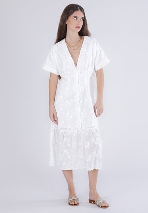 Donna con lunghi capelli scuri che indossa un vestito midi bianco ricamato con fiori e collane dorate a strati, in piedi davanti a uno sfondo chiaro e uniforme.