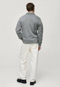 Grå tröja med rundad krage och ribbade ärmslut, kombinerad med vita raka byxor och svarta skor, sedd bakifrån.