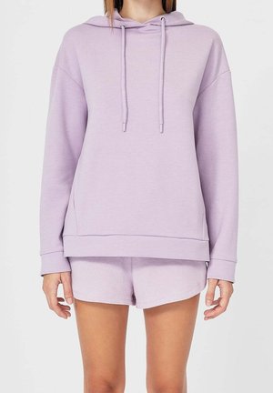 Personne portant un sweat à capuche violet clair et un short assorti, debout devant un fond gris clair uni.