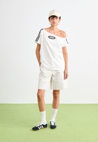 Jauns cilvēks, kurš valkā baltu cepuri, off-shoulder Adidas t-kreklu, svītrainus ceļa garuma šortus, baltas zeķes un melnas Adidas kedas, stāv uz zaļas grīdas.