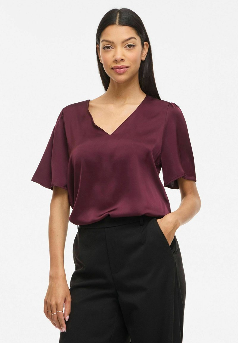 Femme aux cheveux longs et foncés portant un chemisier bordeaux à manches courtes et un pantalon noir, une main dans la poche, debout devant un fond blanc.