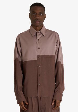 Uomo che indossa una camicia larga bicolore con bottoni, metà superiore marrone chiaro e metà inferiore marrone scuro, pantaloni marrone scuro abbinati.