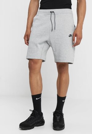 Mann trägt hellgraue Nike-Shorts mit schwarzer Kordel, schwarze Nike-Socken und schwarze Turnschuhe, steht vor einem einfarbigen Hintergrund.