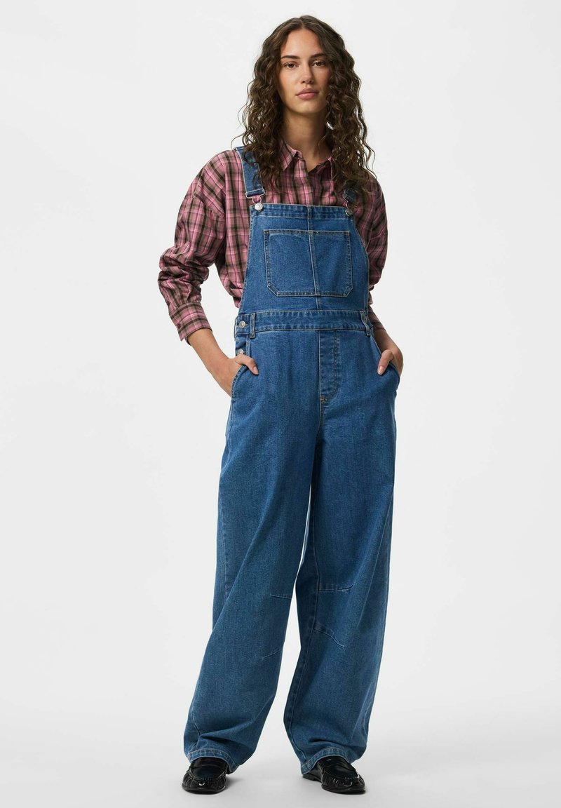 Femme aux cheveux bouclés portant une chemise à carreaux rose et une salopette en denim bleu ample, debout les mains dans les poches.