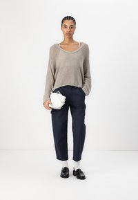Beige gestrickter Pullover, lockere Passform, über dunkelblauen Hosen. Eine kleine weiße Tasche wird in einer Hand gehalten, schwarze Schuhe und weiße Socken vervollständigen das Outfit.
