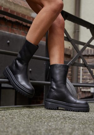 Jambes portant des bottes en cuir noir mi-mollet avec des semelles épaisses et des fermetures éclair, posées contre une rambarde métallique urbaine et un mur en briques en arrière-plan.