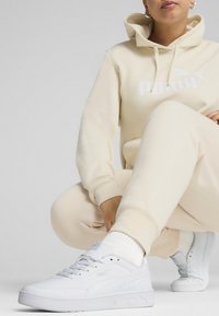 Beige hoodie met wit "PUMA" logo, bijpassende beige broek en witte sneakers met textuurzolen, voorzien van subtiele merkaanduidingen.