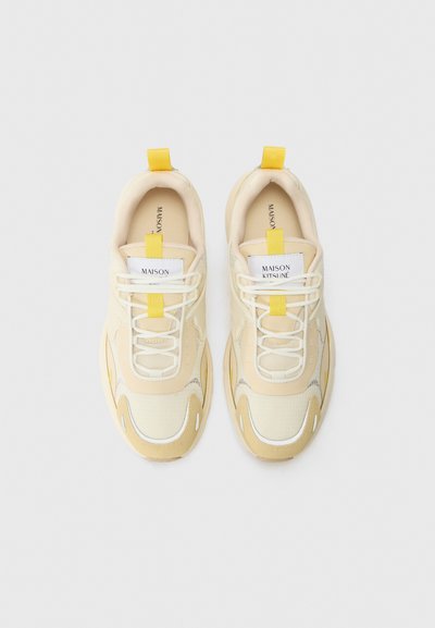 Baskets beiges avec une tige en maille texturée, accents blancs et lacets jaunes. Elles présentent un col rembourré et des étiquettes de logo sur la languette.