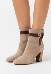 Botas de tobillo de ante beige con puntera afilada, tacones de bloque y correas marrón oscuro con hebillas en tono dorado, llevadas sobre piernas desnudas.
