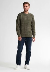 Olivgrüner, grob gestrickter Pullover mit rundem Ausschnitt, aus strukturiertem Stoff, kombiniert mit dunkelblauen Jeans und weißen Turnschuhen.