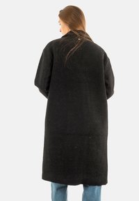Donna con capelli lunghi, che indossa un lungo cappotto nero texturato e jeans blu, in piedi di spalle su uno sfondo neutro.