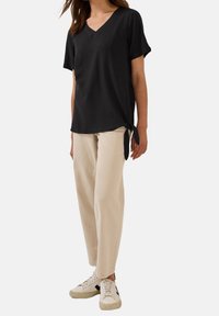 T-shirt nera con scollo a V, maniche corte e dettaglio a nodo laterale, abbinata a pantaloni larghi beige e sneakers bianche con accenti neri.
