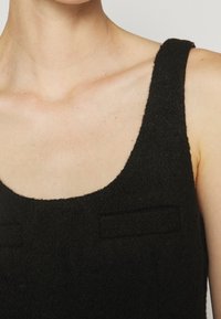 Gros plan du haut du torse portant un top noir sans manches en tissu texturé avec un col arrondi et des fines bretelles.