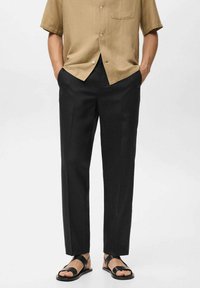 Pantaloni neri con una vestibilità dritta, caratterizzati da una superficie liscia e un leggero lucido. Abbinati a una camicia beige a maniche corte e sandali neri.