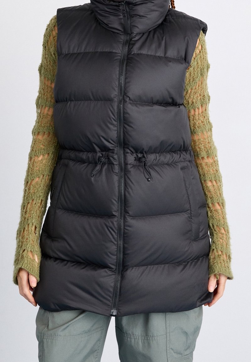 Veste sans manches noire matelassée avec un col haut, fermeture éclair, taille ajustable par cordon de serrage, et sections matelassées verticales. Portée par-dessus un pull en tricot vert.