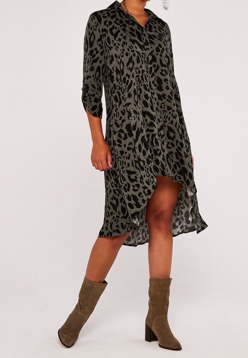 Femme portant une robe high-low à manches longues, imprimée léopard vert olive et noir, manches retroussées et bottines en daim marron.