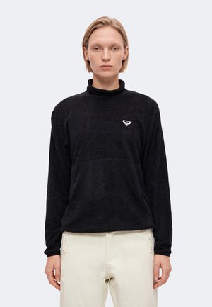 Un mannequin aux cheveux courts et blonds porte un pull noir à col montant et à la texture travaillée, ainsi qu’un pantalon blanc. Il fait face à l’objectif devant un fond uni et clair.