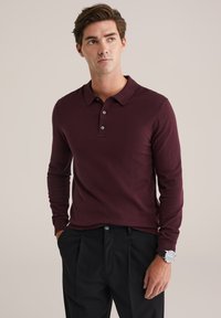 Férfi hosszú ujjú bordó pólót és fekete nadrágot visel, bal csuklóján órával, semleges háttér előtt áll.