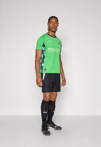 Zelena nogometna majica s črnimi dodatki, kratkimi rokavi in kamuflažnim vzorcem na straneh. Oblečena z rjavimi kratkimi hlačami in dolgimi črnimi nogavicami.