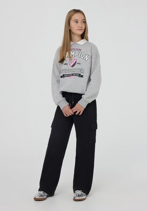 Felpa grigia con stampa "Champion Supporter", abbinata a pantaloni cargo neri con tasche laterali e sneakers argento con finitura lucida.