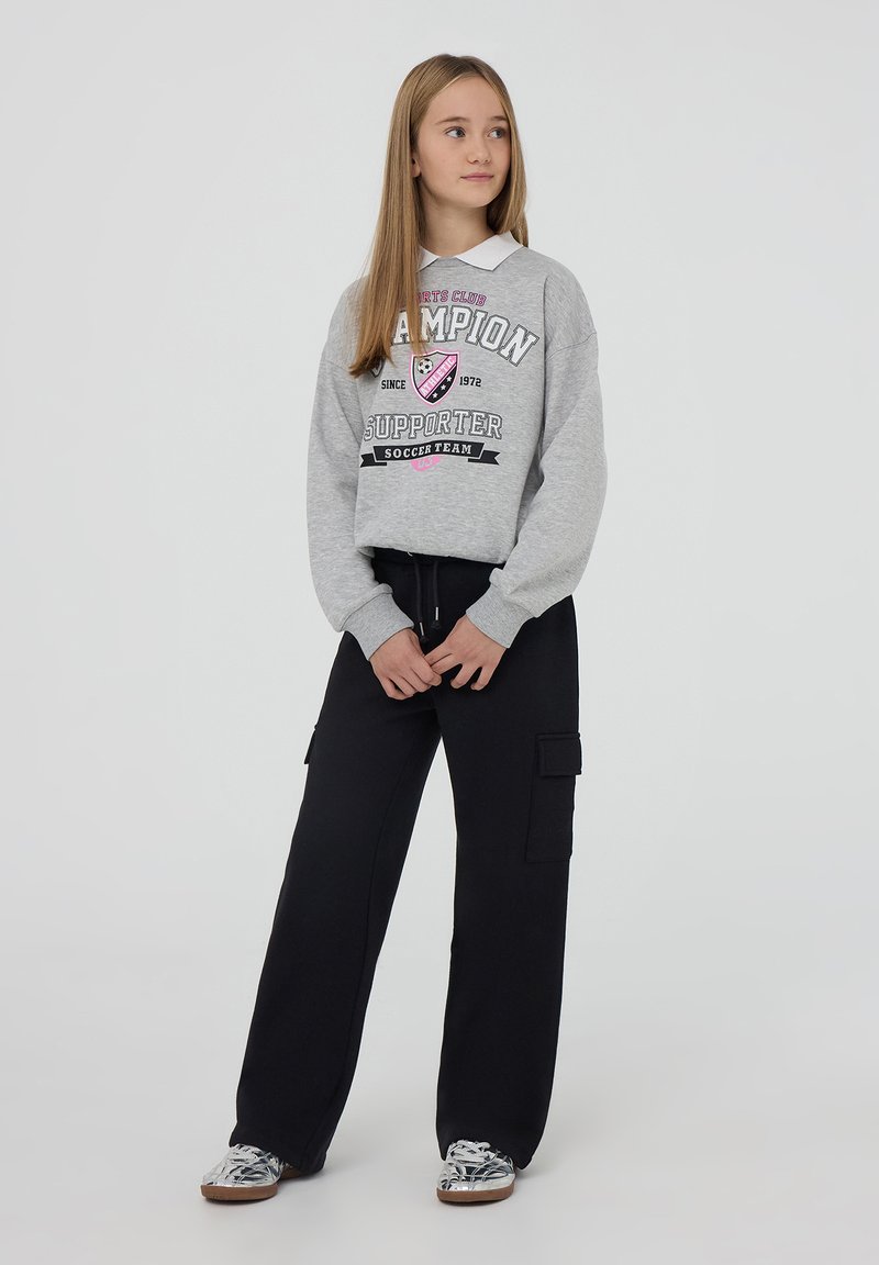 Felpa grigia con stampa "Champion Supporter", abbinata a pantaloni cargo neri con tasche laterali e sneakers argento con finitura lucida.