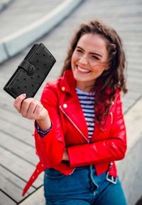 Black leather wallet with a textured, dotted pattern and a round logo clasp, held by a person in a red jacket against a wooden background.