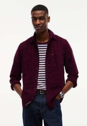 Junger Mann trägt ein burgunderfarbenes Hemd über einem gestreiften T-Shirt, blaue Jeans und eine Uhr, steht mit den Händen in den Taschen.