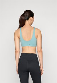 Ženska sportska majica u nijansi tirkizne s širokim trakama i dizajnom za trkače leđa, s suptilnim Nike logom. Uparena s crnim tajicama visokog struka.