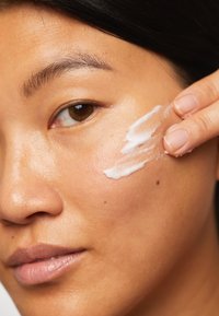 Crème hydratante appliquée sur la joue ; la crème est blanche, avec une texture lisse. La peau semble hydratée, avec des pores visibles et de légères taches de rousseur.