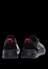 Baskets en cuir noir avec des accents rouges et noirs, des détails en daim, une pointe ronde et une semelle épaisse et texturée. À l'arrière, un rabat contrastant.