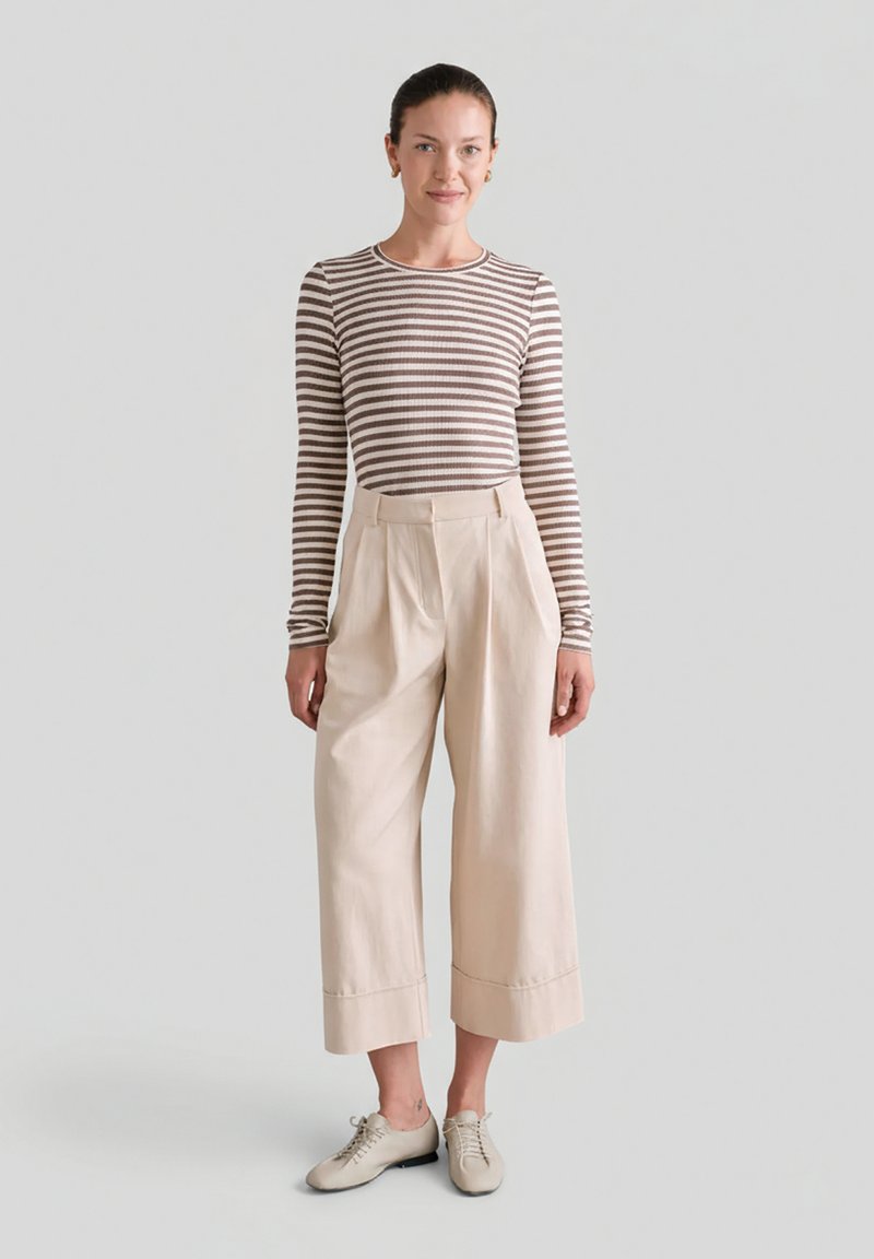 Kobieta stojąca w brązowo-białej, paskowanej bluzce z długim rękawem, beżowych, szerokich spodniach o skróconej długości oraz beżowych sznurowanych butach.