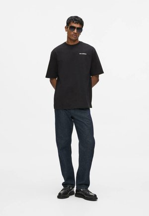 Mężczyzna w czarnej, oversize koszulce z małym, białym logo, ciemnoniebieskich dżinsach, czarnych butach i czarnych okularach przeciwsłonecznych, stojący z rękami za plecami.