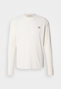 Wit thermoshirt met lange mouwen en geribbelde textuur, voorzien van een klein beige logo-patch op de borst en aansluitende manchetten.