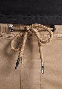 Beige trekkoord met zilveren metalen uiteinden, door metalen oogjes geregen, gecombineerd met een donkere shirtzoom aan de bovenkant, die een zachte stoffen textuur toont.