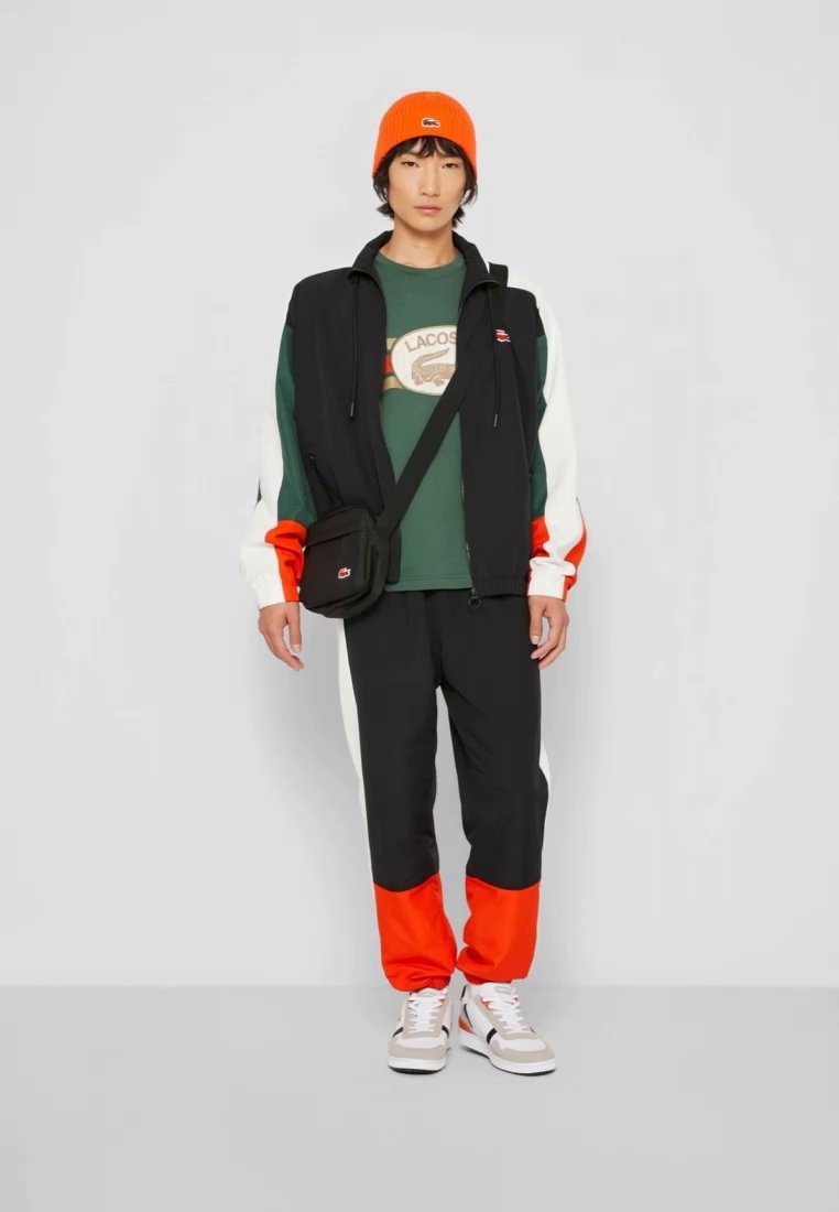 Gilet noir zippé, chemise verte avec logo, pantalon noir avec accents orange, manches blanches, bonnet orange, sac bandoulière, sneakers blancs.