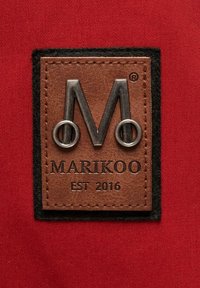 Hnedá kožená nálepka s kovovým logom "M", čiernym zamakom a textom "MARIKOO EST 2016" na červenom textilnom pozadí.