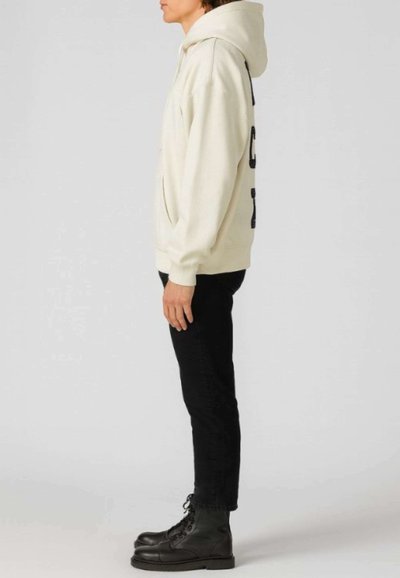 Felpa beige chiaro con tasca frontale, presenta un logo nero su un lato, abbinata a jeans neri slim e stivali neri stringati.