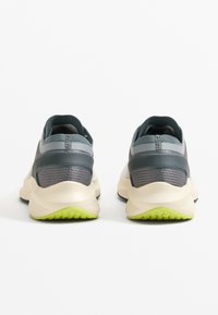 Férfi sportcipők szürke hálós felsőrésszel, zöld gumitalppal, fényvisszaverő részletekkel és vastag, párnázott középtalppal.